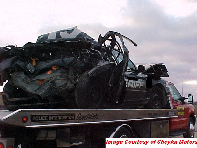 Wrecked black police car with "SHERIFF" text, on a red tow truck. Front end is crushed