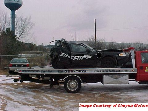 2000 Ford Crown Victoria - A wrecked 2000 Ford Crown Victoria police car is being towed. Another car is parked nearby.