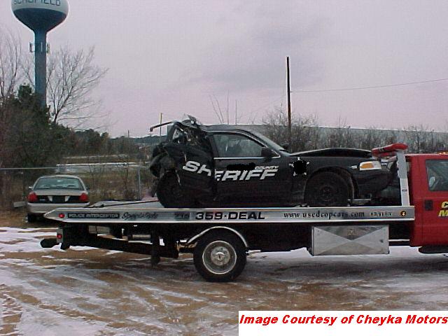 2000 Ford Crown Victoria police car, heavily damaged, loaded on a tow truck. Another car is visible on the ground