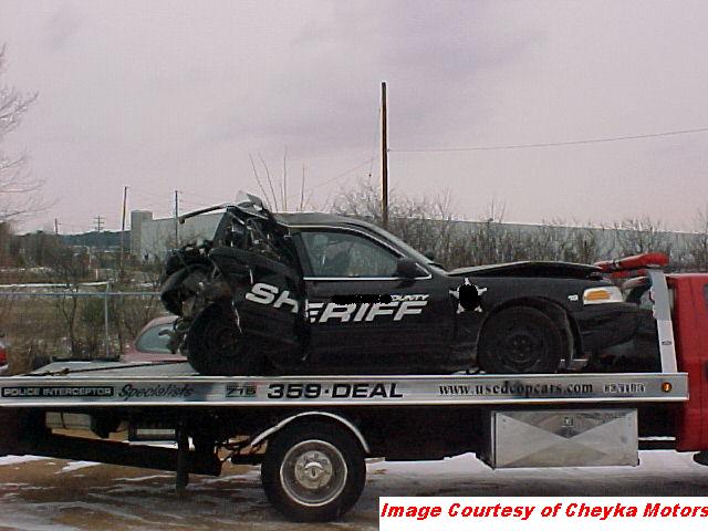 Damaged black sheriff's car with 'SHERIFF' text on its side, loaded onto a tow truck