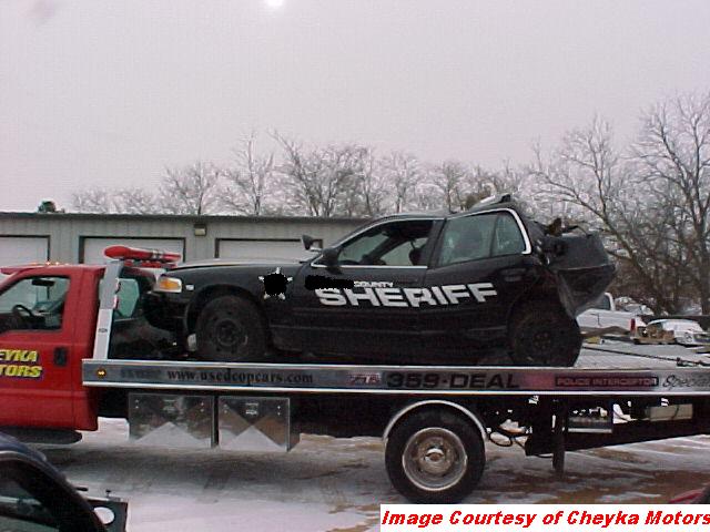Damaged black sheriff's car with 'COUNTY SHERIFF' on the side, being towed on a red truck