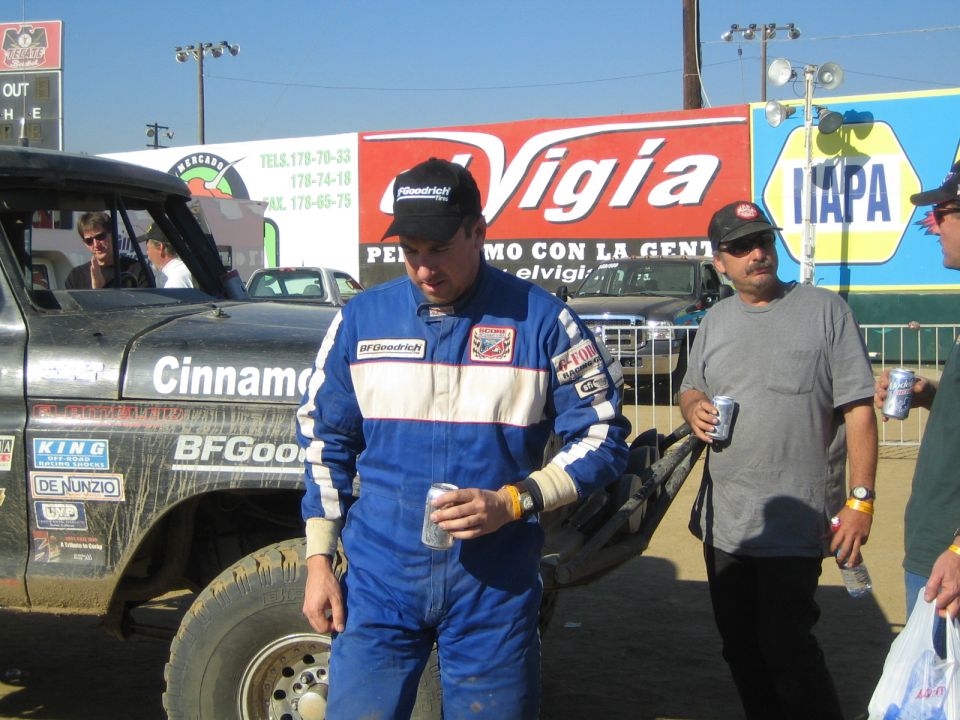 Man in a blue BFGoodrich racing suit holding a can beside a race truck and Vigia banner