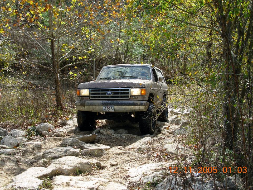 1987 Ford Bronco navigating a rocky off-road path with trees and foliage surrounding it