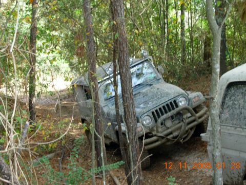 1987 Ford Bronco - A Jeep Wrangler navigates a muddy, wooded off-road trail, with another vehicle visible to the right.