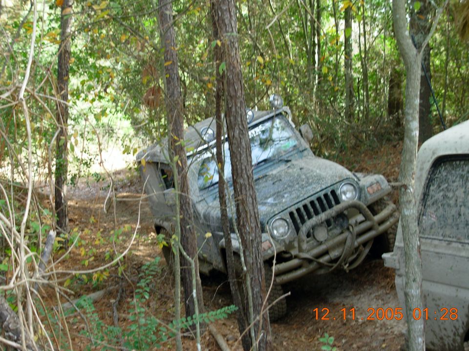 Muddy Jeep Wrangler on wooded trail, another vehicle partially visible
