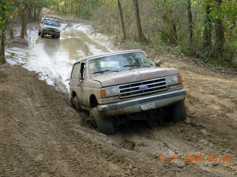 1987 Ford Bronco - A 1987 Ford Bronco navigates a muddy off-road trail, with another vehicle visible in the background.