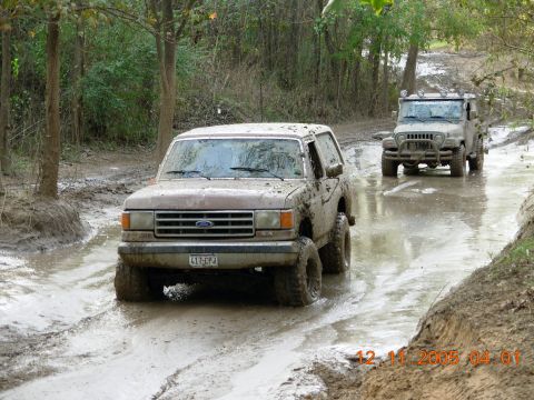 1987 Ford Bronco - Two off-road vehicles navigate a challenging muddy path, kicking up water and dirt.