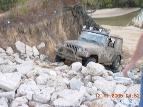 1987 Ford Bronco - A muddy Jeep Wrangler attempts to traverse a difficult, rocky terrain near a riverbank.