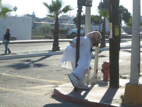 Person in a white suit and bald mask leaning on a street post near a fire hydrant