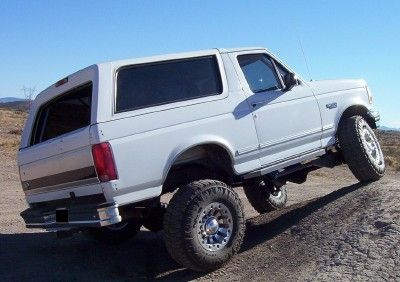 1995 Ford Bronco - A white 1995 Ford Bronco is shown from the rear-left quarter angle, climbing a dirt slope with its suspension lifted.
