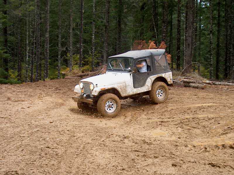 White Jeep CJ-5 with black soft top driving through mud in a forest clearing