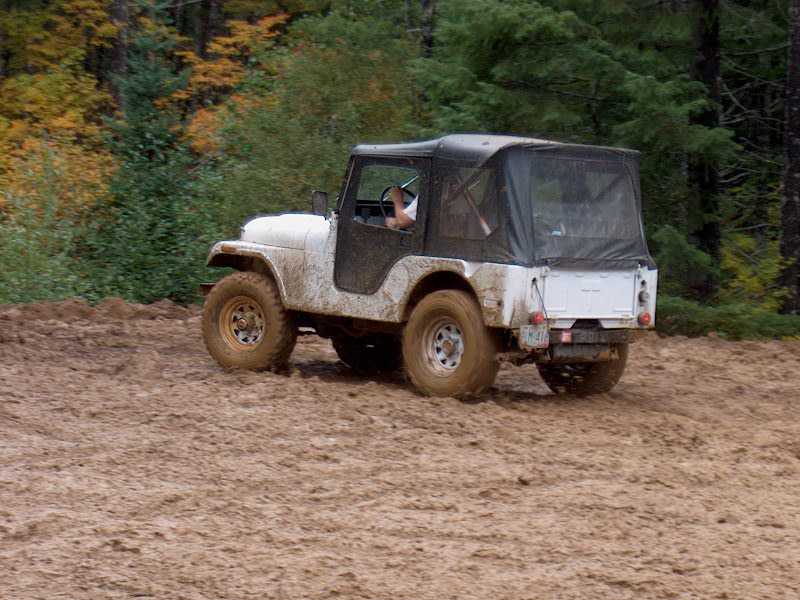 White Jeep CJ-5 with black soft top driving across a muddy off-road field by autumn woods
