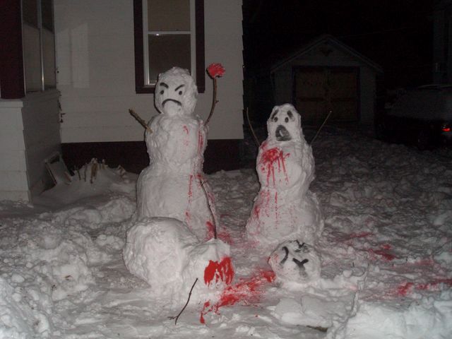 Two snowmen, one with an angry face and one with a scared face, are covered in fake blood. A third snowman head lies on the ground