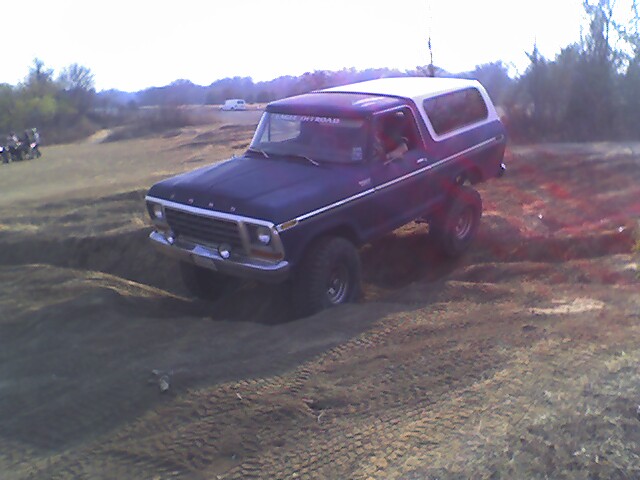 Late-70s Ford Bronco with black hood and burgundy body, white top, on a dirt berm in a grassy field