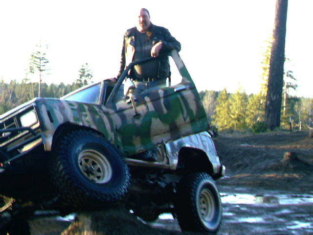 Man standing in a camo-painted compact pickup with doors removed, climbing a muddy off-road obstacle