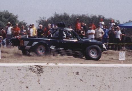 1984 Ford Bronco - A modified pickup truck competes on a dirt track in front of a crowd.