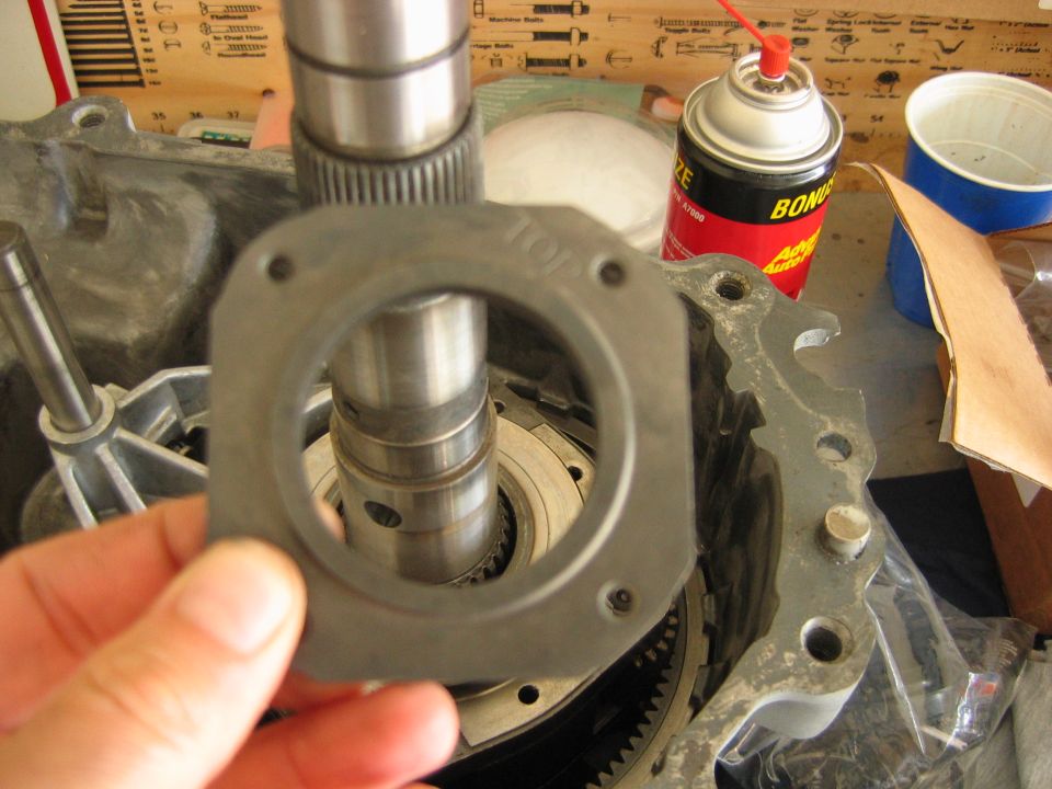 Hand holding a black gasket marked TOP over the output shaft of a disassembled transmission on a workbench