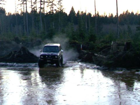 1987 Ford Bronco II - A dark 1987 Ford Bronco II navigates a muddy, water-filled path surrounded by trees.