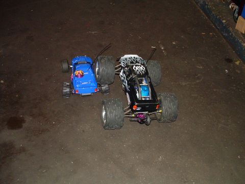 1994 Ford Bronco - Two RC monster trucks, one blue and one black with flame decals, sit on a dark, textured surface.