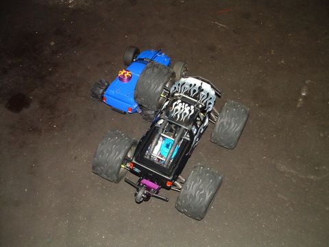1994 Ford Bronco - Two remote control monster trucks are positioned on a dark, possibly wet, surface.