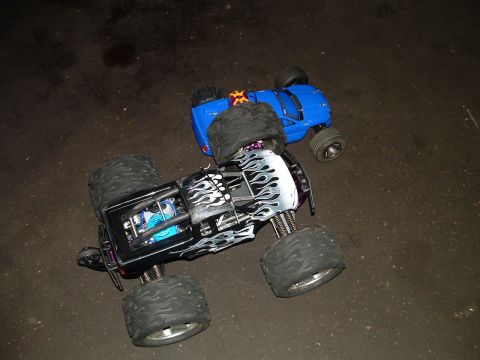 1994 Ford Bronco - Two remote control monster trucks, one black with flame decals and the other blue, are positioned on a dark, possibly asphalt, surface.