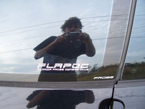 1994 Ford Probe - A man photographs his reflection in the window of a car, which displays club stickers.