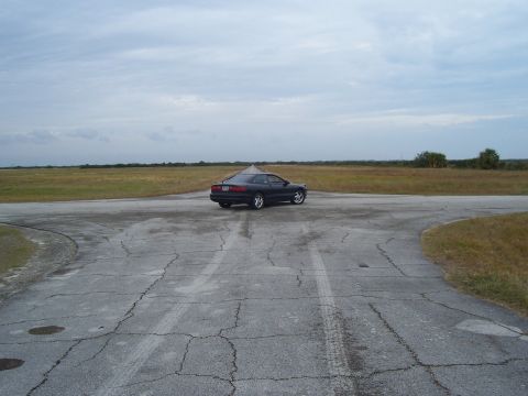1994 Ford Probe - A dark coupe sits on a cracked asphalt road in a wide-open, grassy area with a cloudy sky overhead.