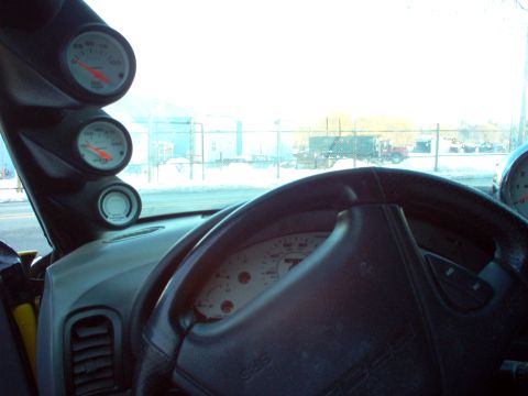 1994 Ford Probe - The interior of a vehicle shows a dashboard with multiple gauges and a steering wheel, with a view of trucks outside.