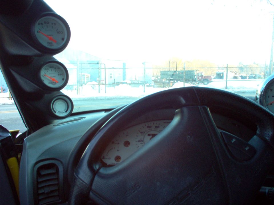Dashboard with three gauges mounted on the A-pillar, steering wheel, and speedometer visible. Outside view shows a fenced area with trucks