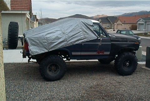 1986 Ford Bronco - A 1986 Ford Bronco is partially covered by a silver tarp, revealing its side profile and a rear-mounted spare tire.