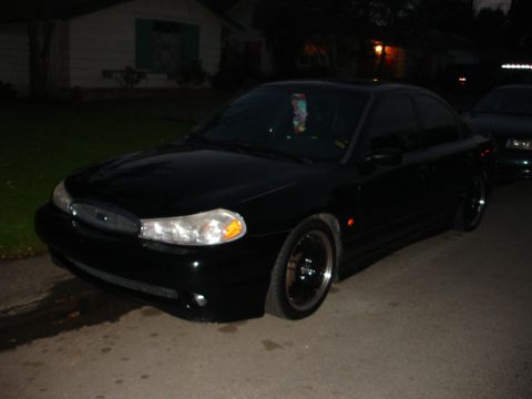1995 Ford Taurus - A black 1995 Ford Taurus sedan is parked on a residential street at night, with another vehicle visible in the background.