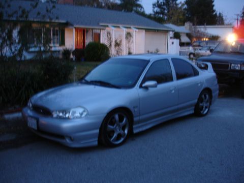 1995 Ford Taurus - A silver sedan with aftermarket wheels is parked in front of a house, with a dark SUV behind it.