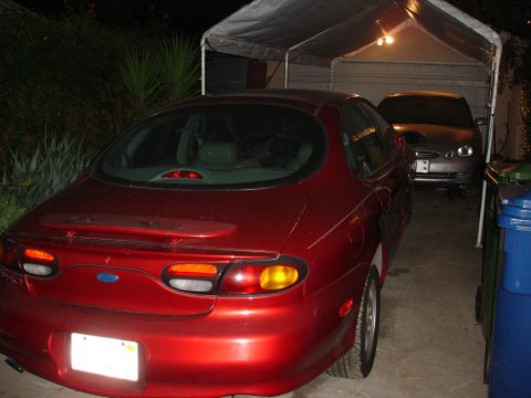 1995 Ford Taurus - A red 1995 Ford Taurus is parked in a carport next to another vehicle.