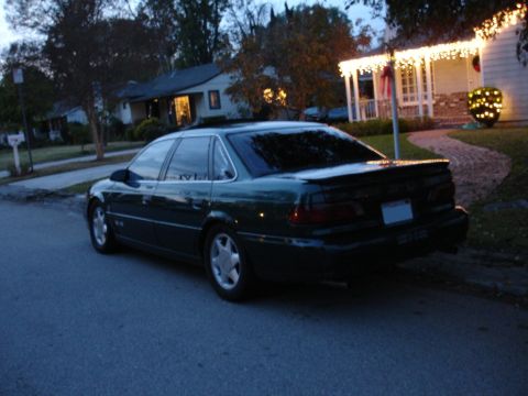 1995 Ford Taurus - A dark green sedan is parked on a residential street in front of a house decorated with Christmas lights.