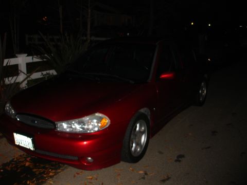 1995 Ford Taurus - A red 1995 Ford Taurus sedan is parked on a street at night, showing its front passenger side and license plate.