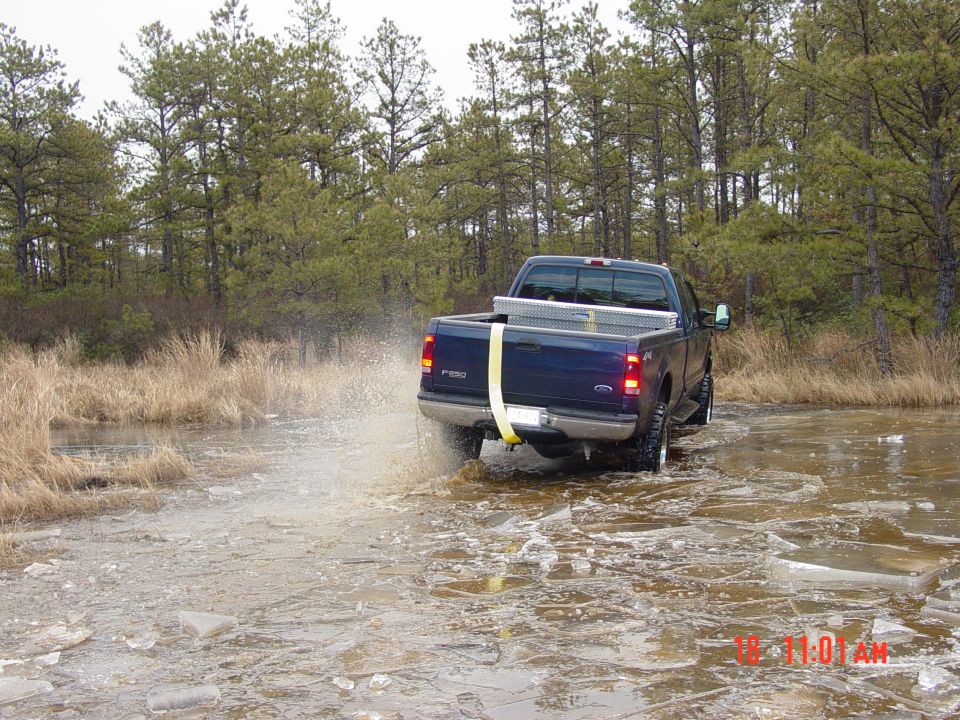 Blue pickup truck with a toolbox in the bed drives through shallow water, kicking up spray. The truck has "F-250" badging