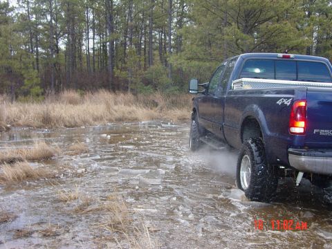 1995 Dodge 1500 - A blue pickup truck navigates through a shallow body of water covered in ice and debris, kicking up spray as it moves.
