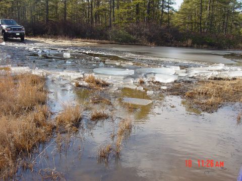 1995 Dodge 1500 - A dark pickup truck navigates a road covered in ice and broken ice chunks, with a wooded area visible behind it.
