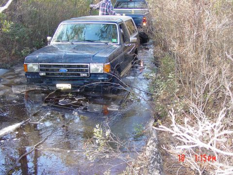 1995 Dodge 1500 - A dark blue Ford Bronco is partially submerged in water, with a pickup truck following behind it on a wet path.