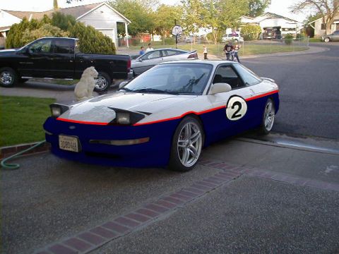 1997 Ford Probe - A two-tone sports car with a racing number is parked in a driveway next to a pickup truck.