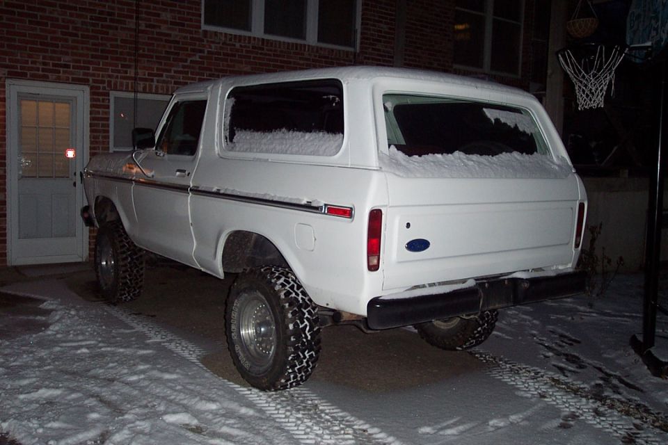 White lifted Ford Bronco SUV from rear at night on snowy driveway by brick wall