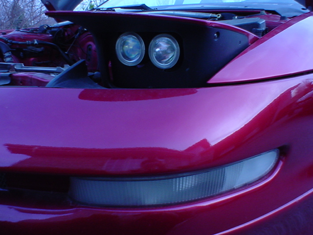 Close-up of a red sports car's front end, showing pop-up headlights and a corner marker light