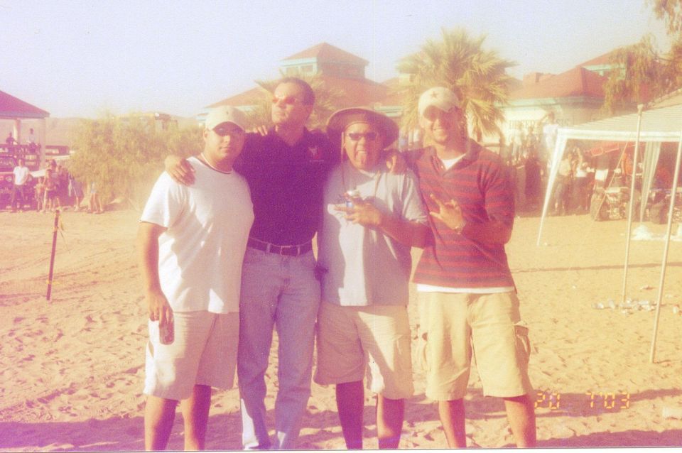 Four men stand together on a sandy beach with buildings and palm trees in the background