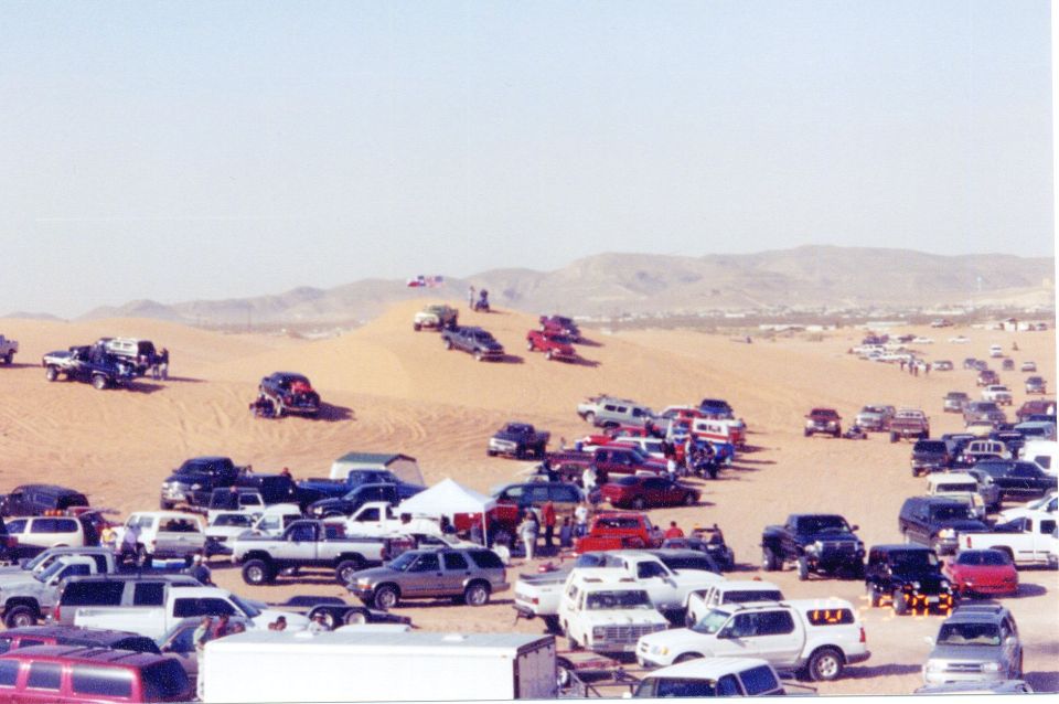 Sandy desert staging area packed with rows of parked trucks and SUVs below dunes under clear sky