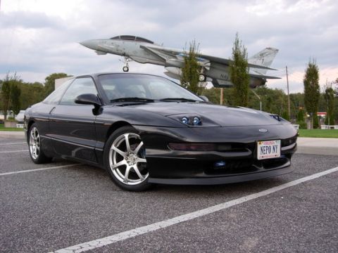 1993 Ford Probe - A black 1993 Ford Probe is parked in front of a fighter jet on a cloudy day.