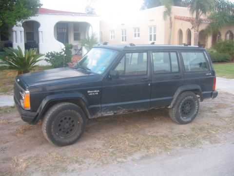 1989 Ford Bronco - A dark colored Jeep Cherokee 4x4 is parked on a dirt lot in front of a house with Spanish-style architecture.