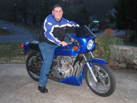 1985 Ford LTD Crown Victoria - A man in a blue and black motorcycle jacket sits astride a blue Suzuki standard/cafe-style motorcycle with a fairing and windscreen, shown…