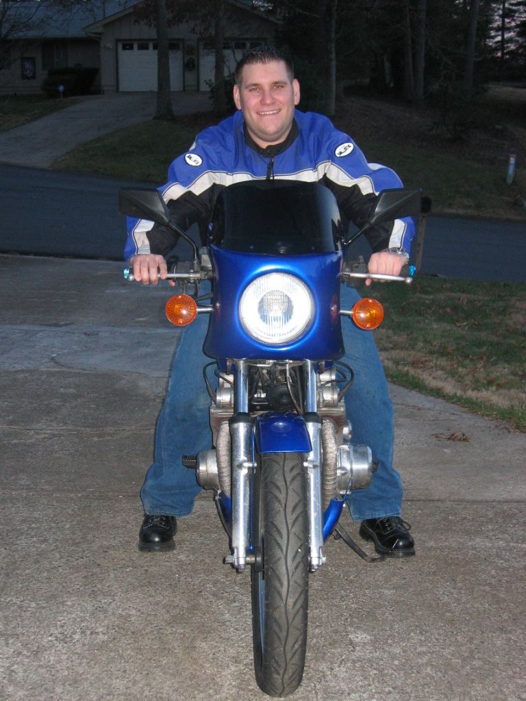 Man in blue jacket on a blue motorcycle, front view. Motorcycle has a round headlight and orange turn signals