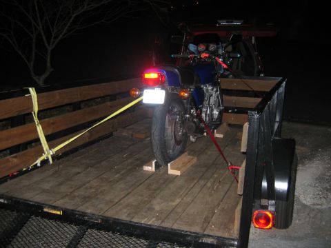 1985 Ford LTD Crown Victoria - A blue motorcycle is tied down in a trailer at night, ready for transport.