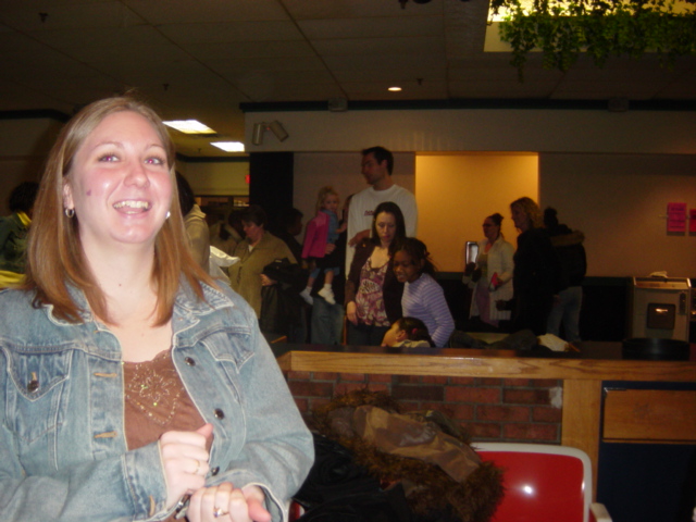 Woman in denim jacket smiling, with a group of people in the background at an indoor event